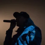 A silhouetted person in a cap and patterned jacket sings into a microphone.