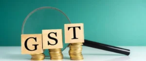 Wooden blocks spelling "GST" are stacked on coins next to a magnifying glass.