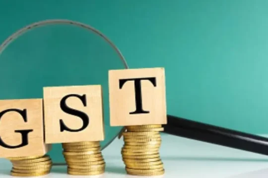 Wooden blocks spelling "GST" are stacked on coins next to a magnifying glass.