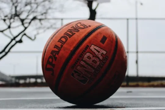 A Spalding NBA basketball rests on a wet outdoor court with a hoop visible.