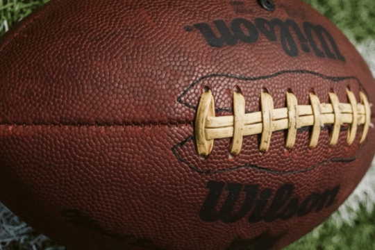 A close-up of a brown Wilson American football resting on green grass with white lines.