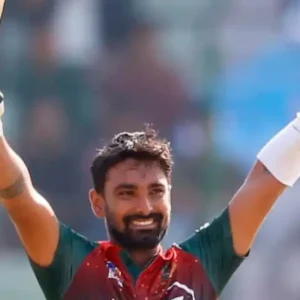 A male cricketer with a beard, wearing a green and red jersey, smiling and raising his bat and helmet in celebration on a cricket field.