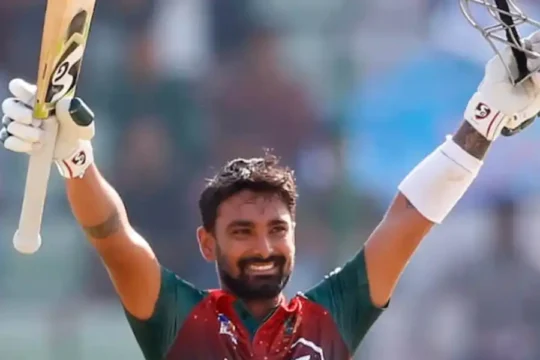 A male cricketer with a beard, wearing a green and red jersey, smiling and raising his bat and helmet in celebration on a cricket field.