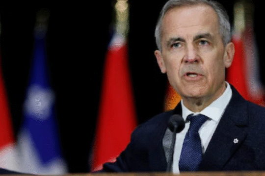 A man in a suit speaking at a podium with flags behind him.