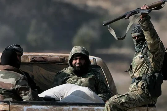 Militiamen in camouflage, one holding a rifle, riding in the back of a pickup truck.