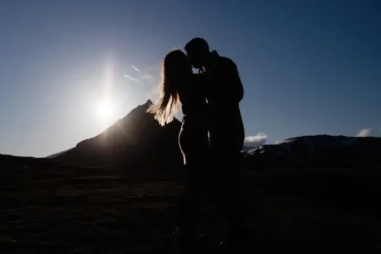 A couple silhouetted against a setting sun behind a mountain.