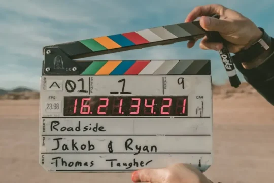 Hands hold a film clapperboard with production details against a desert background.