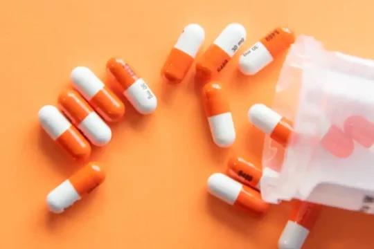Medication capsules, orange and white, scattered from a clear plastic medicine bottle.