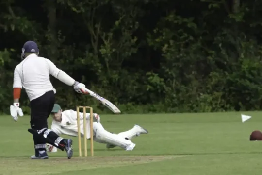A cricket batsman walks away from the wicket, while the wicketkeeper dives.