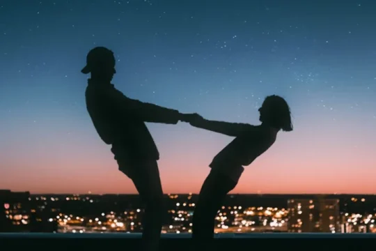 Silhouettes of two people holding hands, leaning back against a city skyline at dusk.