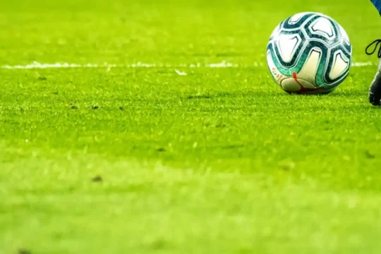 A close-up of a soccer ball and a player's foot in black cleats on a green field.