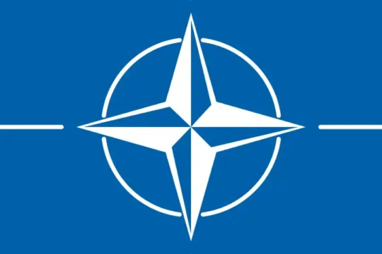 The NATO flag, featuring a white compass rose emblem on a dark blue field with four white lines emanating horizontally.