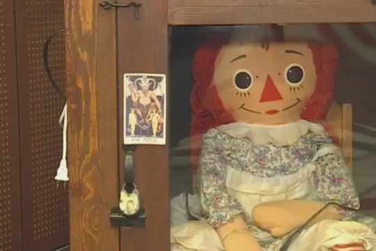 The Annabelle doll is encased in a wooden display cabinet with a tarot card taped to the door.