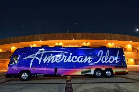 The American Idol tour bus, painted with a starry night sky and purple-to-orange gradient, parked in front of a circular building illuminated at night.