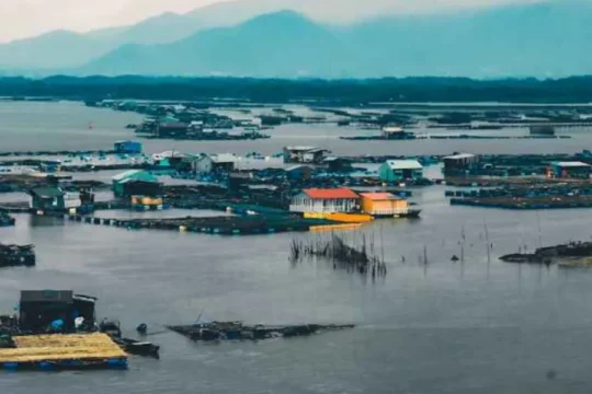 A floating village with numerous houses on a large body of water, mountains in background.