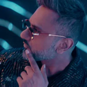 A man with a beard and sunglasses looks away, touching his chin.