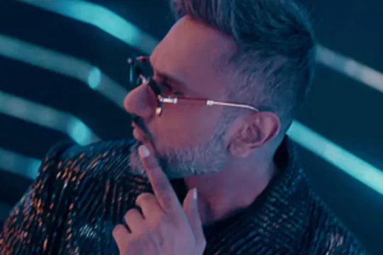 A man with a beard and sunglasses looks away, touching his chin.