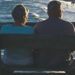 An elderly couple sits on a bench, looking out at the ocean and beach.