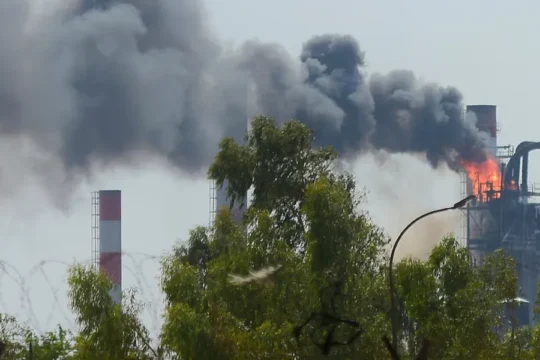 A factory with multiple smokestacks emits black smoke and flames into the sky.