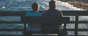 An elderly couple sits on a bench, looking out at the ocean and beach.