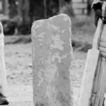 Two children's legs in cricket pads stand beside a bat and concrete block.