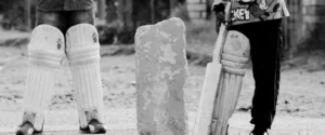 Two children's legs in cricket pads stand beside a bat and concrete block.