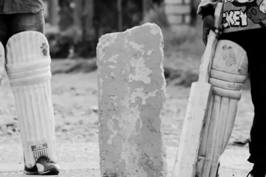 Two children's legs in cricket pads stand beside a bat and concrete block.