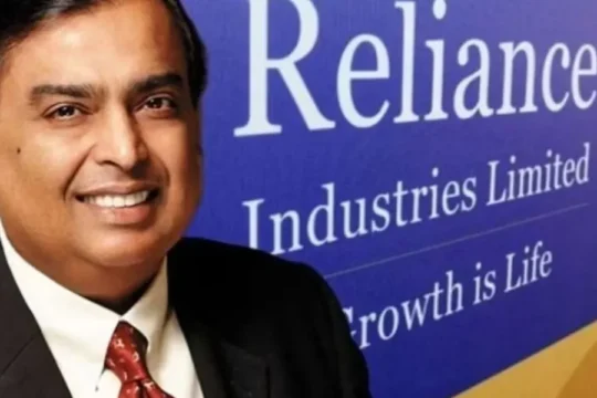 Mukesh Ambani smiles in a suit and tie, with the Reliance Industries logo.