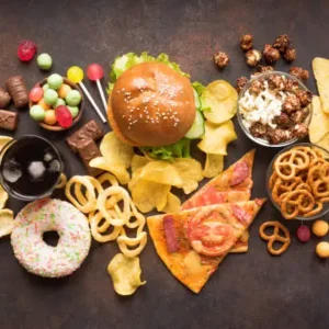 Assorted junk food including burger, chips, pizza, pretzels, candies, and soda on a dark surface.