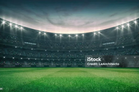 A brightly lit cricket stadium with green grass and a large audience under a cloudy sky at dusk.