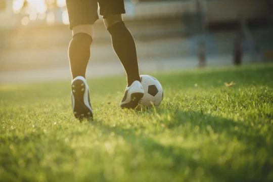 A close-up, low-angle shot shows a soccer player's legs in black shorts, dark socks, and cleated shoes, dribbling a black and white soccer ball across a green grassy field during golden hour with stadium steps in the blurred background.