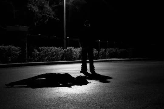 Silhouette of a person standing over a body on a dimly lit road at night, symbolizing a crime or murder scene.