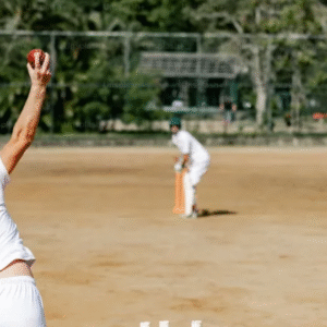 Cricketer bowling a red ball on a dirt pitch with a batsman ready to strike.