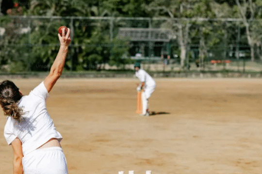 Cricketer bowling a red ball on a dirt pitch with a batsman ready to strike.