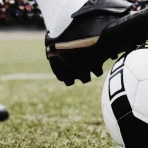 Close-up of a soccer player's cleats with one foot on a black-and-white football on a grassy field.