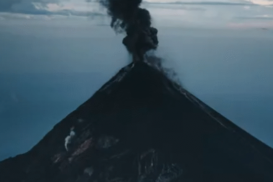 A dark volcanic mountain emits thick black smoke into the sky during an active eruption, surrounded by a hazy blue atmosphere.