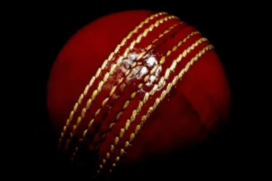 Close-up view of a red leather cricket ball with prominent golden seam stitches against a black background.
