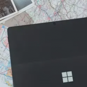 A black Microsoft Surface device placed on a folded map with sunglasses and travel photos nearby.