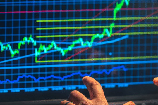 Close-up of a stock market chart on a computer screen showing rising trends with technical indicators, while a person analyzes data using their fingers.