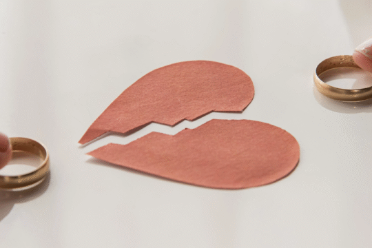 Two hands holding wedding rings beside a torn paper heart symbolizing a breakup or divorce