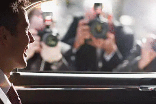 Man smiling in car as paparazzi flash cameras