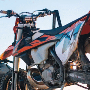 Two off-road motorcycles with helmets parked under a clear blue sky, showcasing rugged tires and motocross graphics.