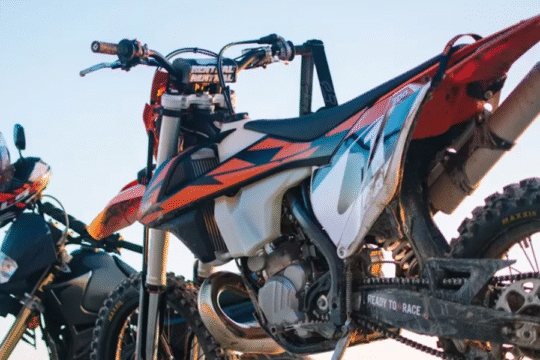Two off-road motorcycles with helmets parked under a clear blue sky, showcasing rugged tires and motocross graphics.