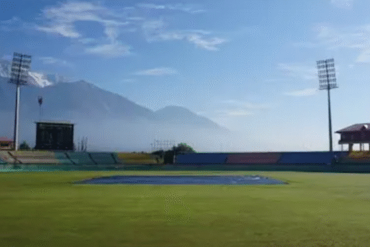 Scenic cricket stadium with snow-capped mountains in the background.