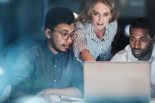 Three professionals working late at night, focused on laptop screen.