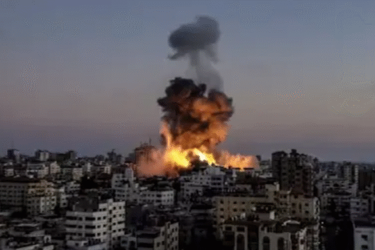 Thick smoke and flames rise from buildings after an airstrike in a densely populated urban area at dusk.