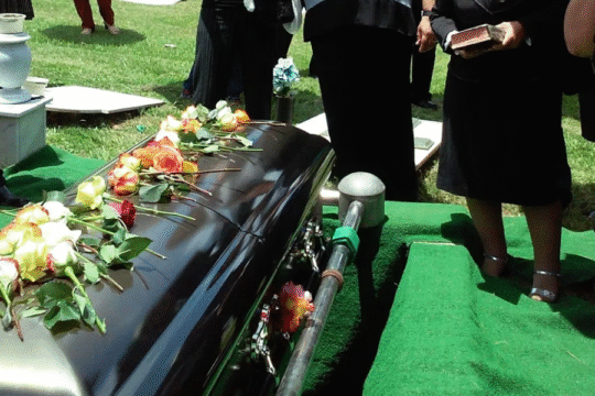 Casket adorned with flowers during a funeral ceremony with mourners in black.