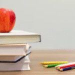 Stack of books with a red apple on top, colorful pencils, and ABC blocks on a wooden table.