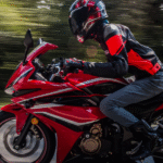 A motorcyclist in red riding gear speeding on a red sports bike on a highway, with trees blurred in the background.