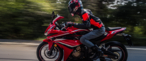 A motorcyclist in red riding gear speeding on a red sports bike on a highway, with trees blurred in the background.